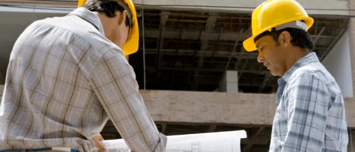 Two men on a construction site, both wearing yellow hard hats and looking at a blueprint