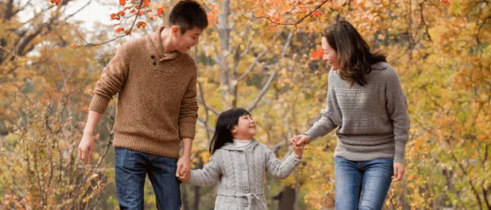 A mother, father, and young daughter walk hand in hand outside, surrounded by fall leaves